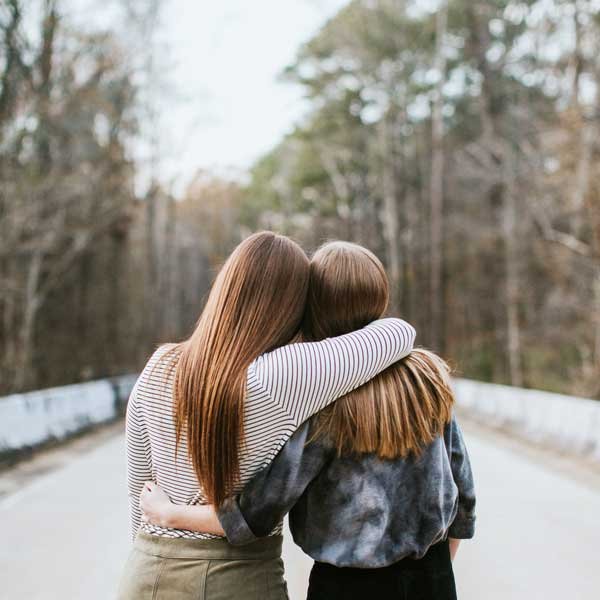 Medium shot of two young ladies hold each other in embrace facing away from the viewer. They are leading into each other and walking along a path.
