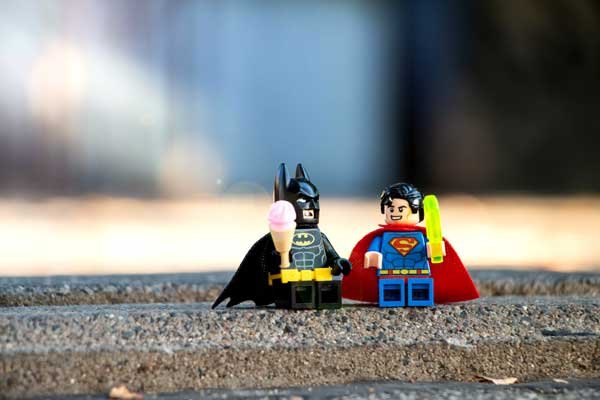 Batman and Superman as Lego figurines sit on a street curb eating ice cream.